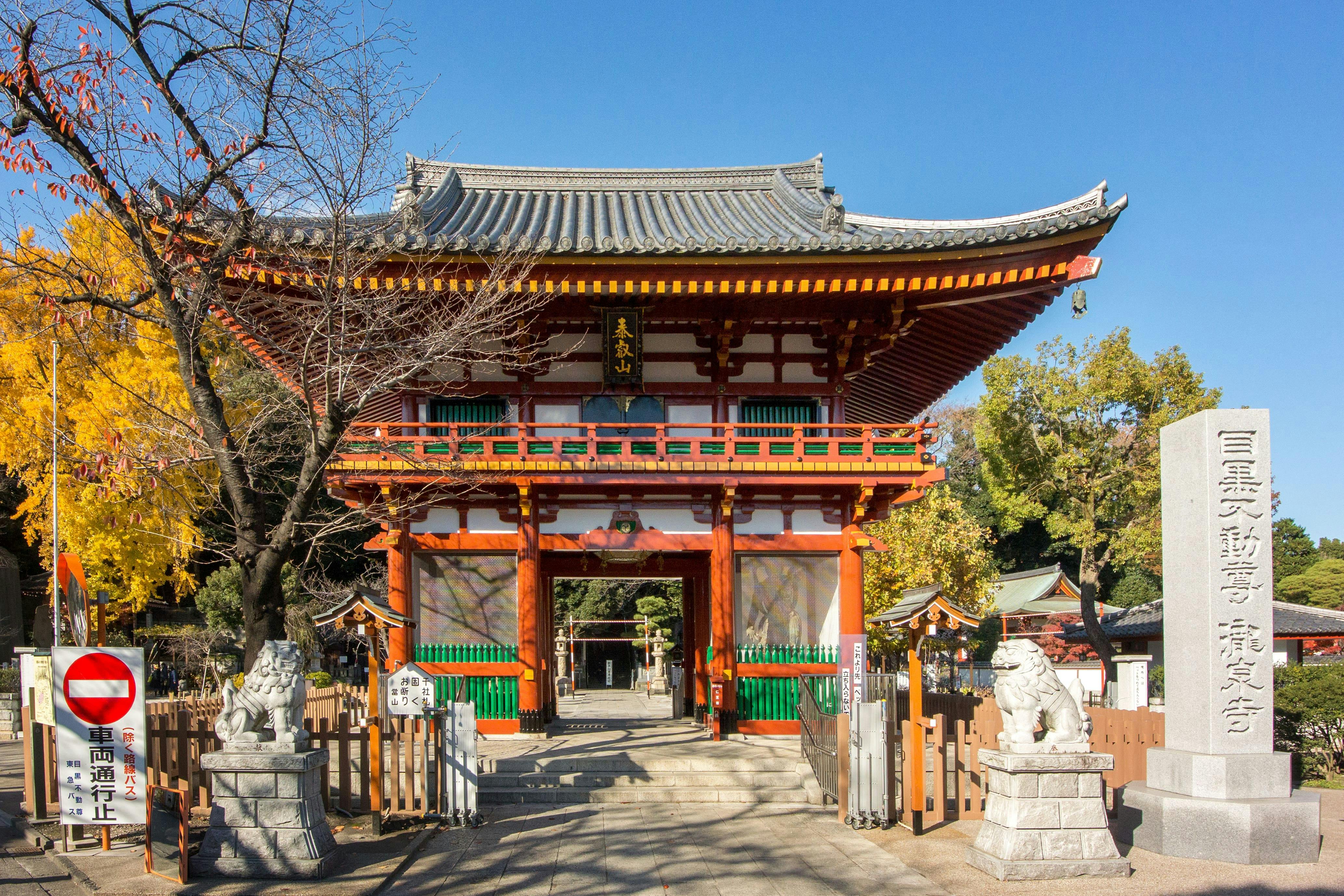 A traditional Japanese temple gate with red pillars and ornate decorations stands under a clear blue sky, flanked by two stone lion statues and surrounded by trees with autumn foliage.