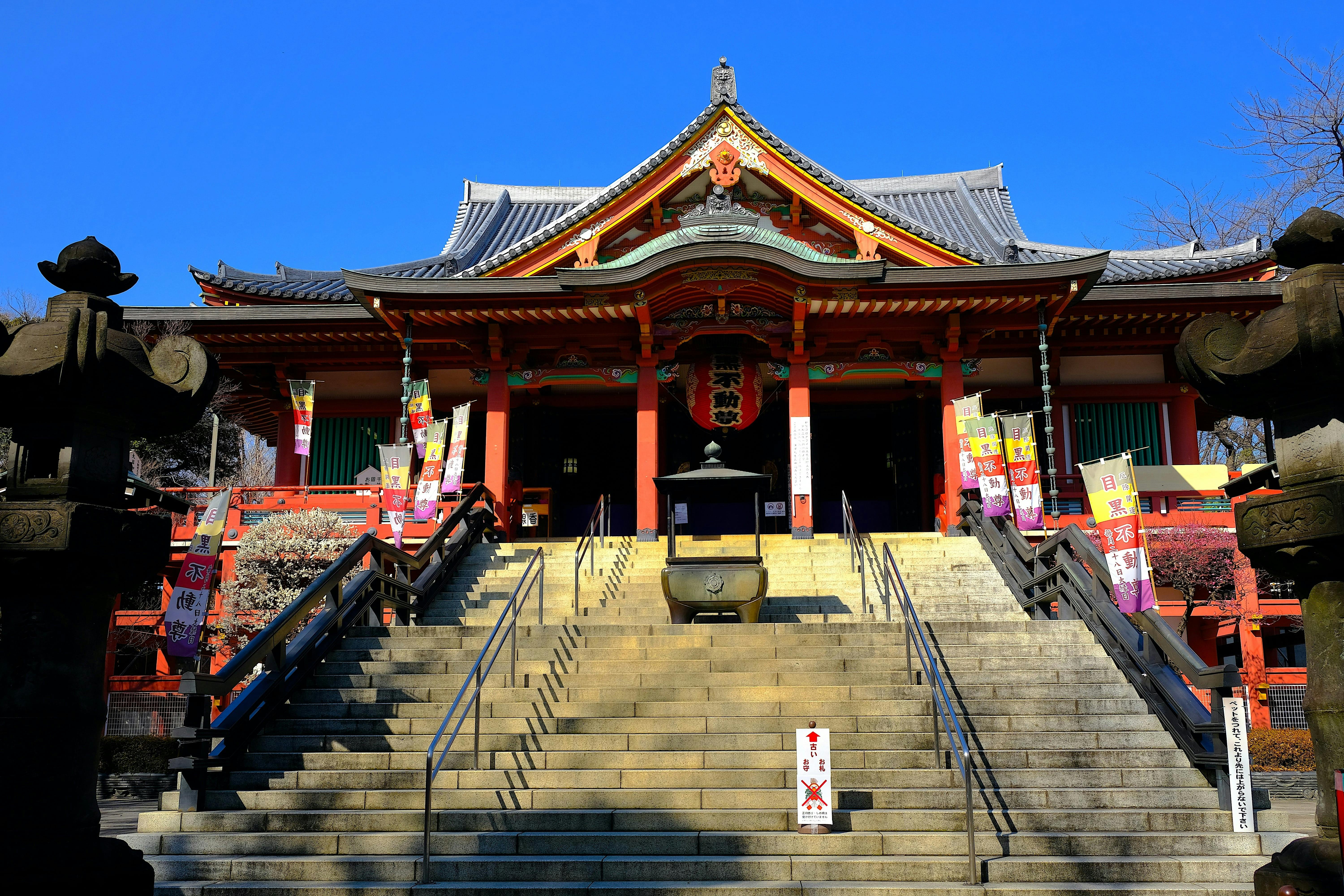 A traditional Japanese temple with ornate roof, colorful banners, and stone lanterns stands atop a wide staircase under a clear blue sky.