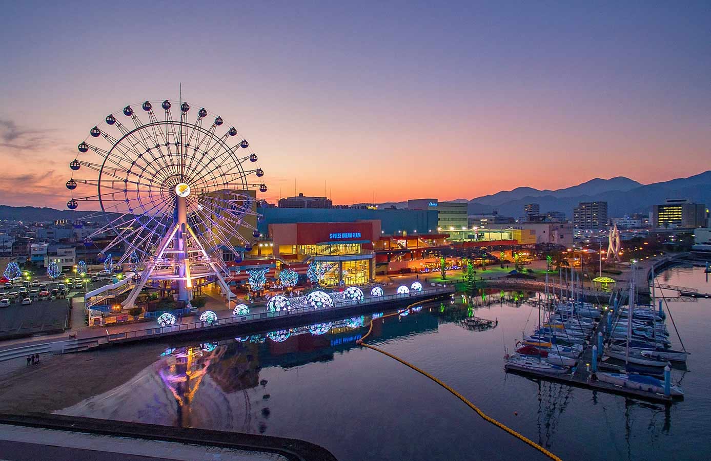 A waterfront cityscape at sunset with a large, illuminated Ferris wheel, colorful buildings, docked sailboats, and reflections on the calm water. Mountains are visible in the background.