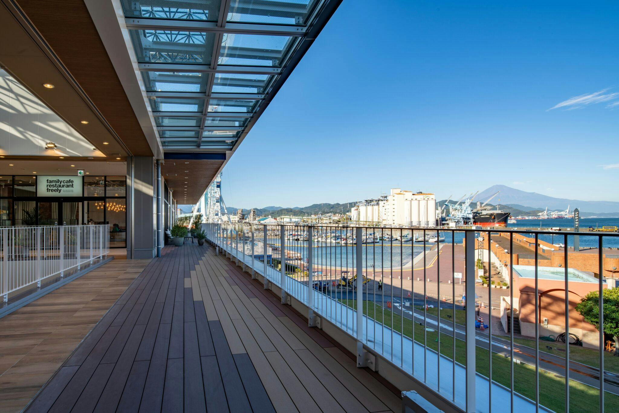 A spacious outdoor terrace with glass railings overlooks a busy harbor, ships, and distant mountains under a clear blue sky. A modern building with large windows and wood flooring lines the left side.