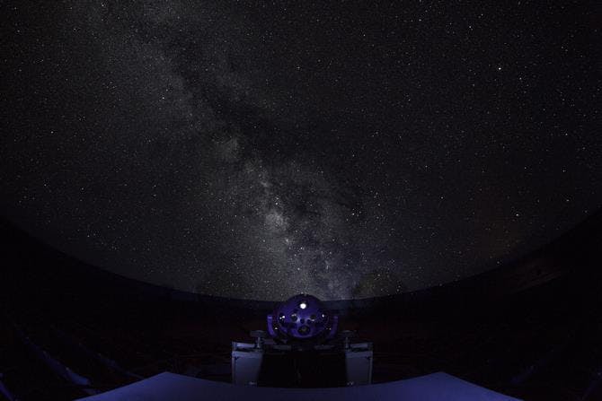 A night sky filled with countless stars and the Milky Way galaxy, viewed from inside a dark observatory dome with a silhouetted planetarium projector in the foreground.