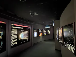 A dimly lit museum hallway displays illuminated panels with astronomical diagrams, images of galaxies, and Japanese text explaining concepts about the universe. The curved wall enhances the futuristic atmosphere.