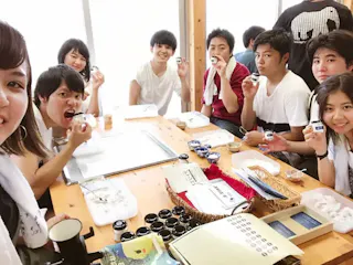 A group of young people sits around a wooden table, smiling and holding small rice balls. Various containers and food items are on the table, and everyone appears to be enjoying a meal together in a bright room.