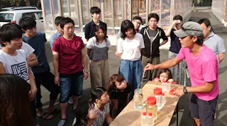 A group of people gathers around a table outdoors, listening to a person in a pink shirt and cap explaining something using jars and bottles filled with liquid. Some are standing, others are sitting, all are focused on the demonstration.