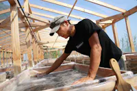 A person in a black t-shirt and cap works with their hands in a shallow, clear water tank inside a wooden structure with a transparent roof, possibly involved in traditional food or craft production.