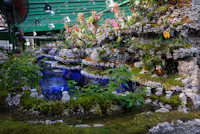 Indoor botanical garden with rocky ledges, lush green plants, and small flowers surrounding a blue-lit pond under a green roof with visible lights and fans.