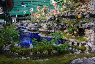 Indoor botanical garden with rocky ledges, lush green plants, and small flowers surrounding a blue-lit pond under a green roof with visible lights and fans.