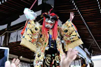 A performer in a traditional Japanese costume and lion mask stands with arms raised on a stage, surrounded by reaching hands and festive decorations.