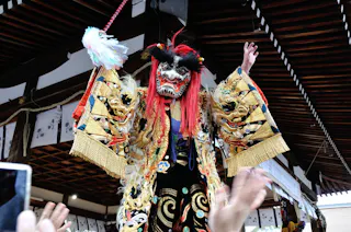 A performer in a traditional Japanese costume and lion mask stands with arms raised on a stage, surrounded by reaching hands and festive decorations.