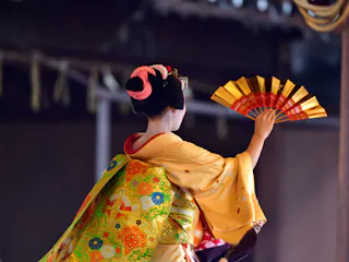 A person in traditional Japanese attire, with an ornate kimono and hair decorations, holds a gold and red folding fan while performing, facing away from the camera against a dimly lit background.