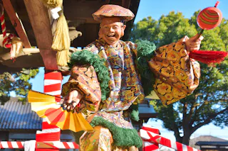 A performer in a colorful, ornate costume and mask holds a decorated fan and stick, smiling during a traditional Japanese festival outdoors, with trees and festive decorations in the background.