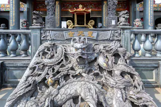 A detailed stone carving of a dragon at the entrance of a traditional Chinese temple, with ornate columns, statues, and golden Chinese characters above the entrance in the background.
