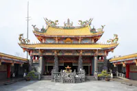 A traditional Chinese temple with ornate, colorful carvings, dragon sculptures on the roof, and intricate details. The temple has a yellow tiled roof and decorative columns, with a courtyard and potted plants in front.