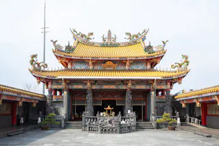 A traditional Chinese temple with ornate, colorful carvings, dragon sculptures on the roof, and intricate details. The temple has a yellow tiled roof and decorative columns, with a courtyard and potted plants in front.