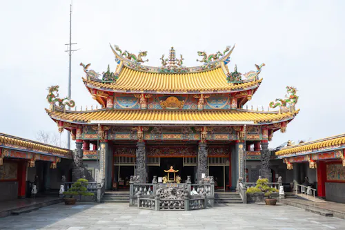 A traditional Chinese temple with ornate, colorful carvings, dragon sculptures on the roof, and intricate details. The temple has a yellow tiled roof and decorative columns, with a courtyard and potted plants in front.