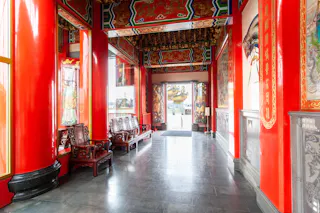 A bright hallway in a Chinese temple features ornate red columns, intricate gold designs on the ceiling, traditional wooden chairs, and open doors leading outside.