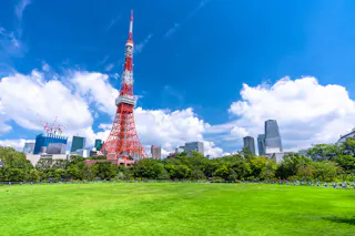 Tokyo Tower stands tall against a bright blue sky with white clouds, surrounded by modern buildings and lush green trees in the foreground, overlooking a large grassy field.