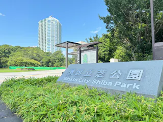 Large stone sign reading "Minato City Shiba Park" in English and Japanese sits among green grass and bushes, with a tall building, trees, and a bright blue sky in the background.