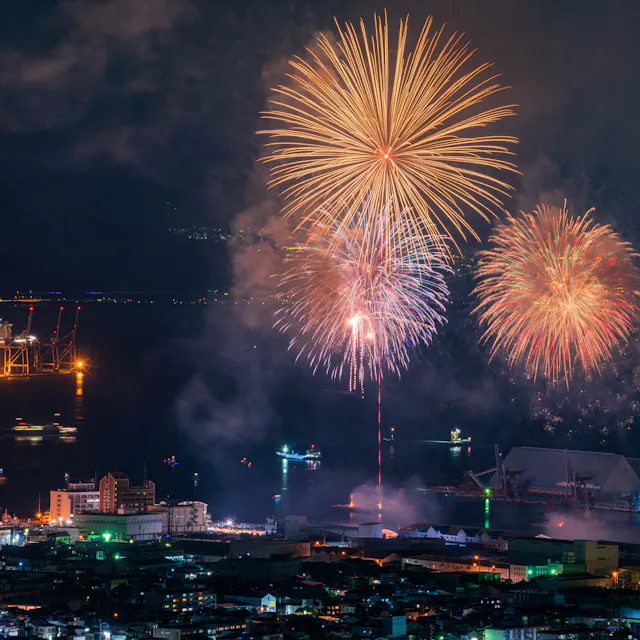 Shimizu Minato Festival Fireworks light up the night sky over a city by the water, with ships and cranes illuminated in the background and city buildings in the foreground.