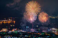Fireworks light up the night sky over a city by the water, with ships and cranes illuminated in the background and city buildings in the foreground.