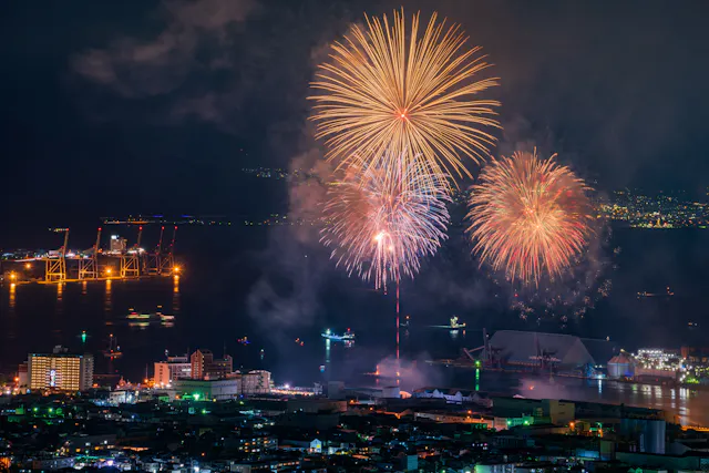 Fireworks light up the night sky over a city by the water, with ships and cranes illuminated in the background and city buildings in the foreground.