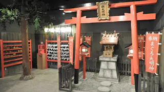 A small indoor Shinto shrine display with red torii gates, traditional lanterns, wooden prayer plaques, paper fortunes tied on strings, and a miniature shrine building, all set on a gravel floor with a tree.