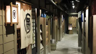 A narrow hallway resembling a traditional Japanese street, with wooden walls, fabric banners, lanterns, and entrances to small shops or restaurants. Japanese characters are visible on signs and curtains. The scene is softly lit.