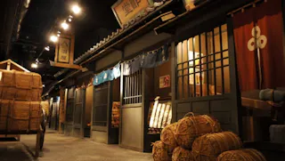 A dimly lit indoor exhibit recreates a traditional Japanese street scene with wooden storefronts, sliding doors, hanging signs, blue noren curtains, and large bundles of straw stacked along the walkway.