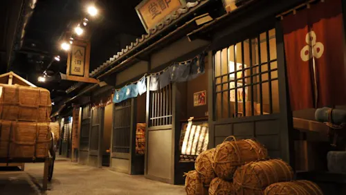 A dimly lit indoor exhibit recreates a traditional Japanese street scene with wooden storefronts, sliding doors, hanging signs, blue noren curtains, and large bundles of straw stacked along the walkway.