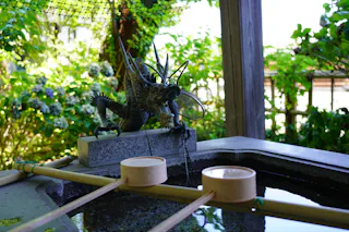 A dragon-shaped water fountain at a Japanese shrine purification basin, with wooden ladles resting on the basin and lush greenery in the background.