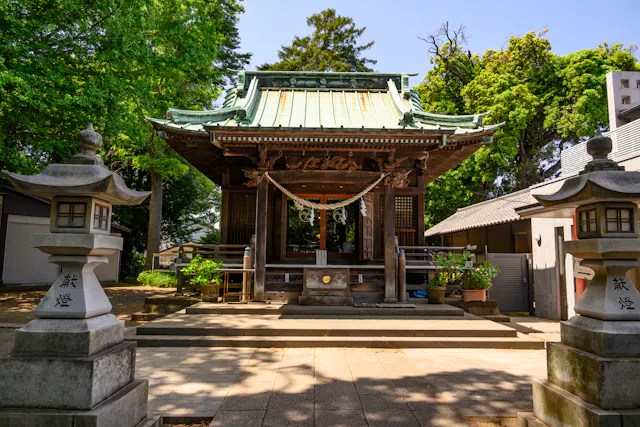 A traditional Japanese Shinto shrine with a green roof, wooden structure, and decorative elements, flanked by two stone lanterns, surrounded by trees on a sunny day.