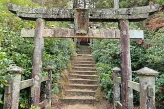 A weathered stone torii gate stands before moss-covered steps leading up through lush greenery to a traditional Japanese shrine in a forested area.
