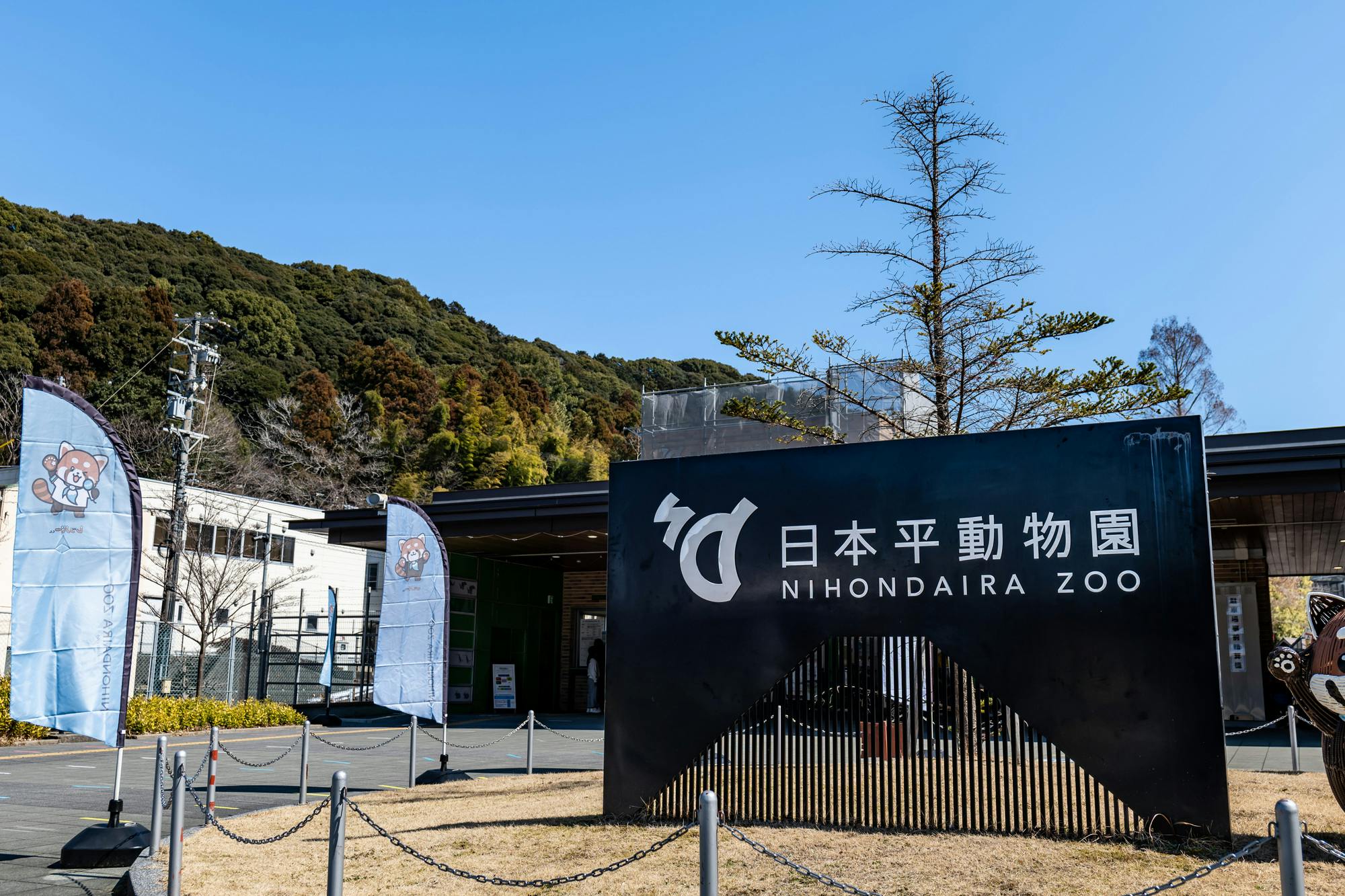 Large black sign reading "Nihondaira Zoo" in English and Japanese stands at the entrance, with banners featuring animal illustrations nearby. Trees and a building are visible in the background under a clear blue sky.
