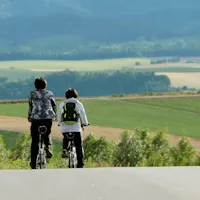 Riding toward the patchwork hills of Biei in summer. Two cyclists riding side by side up a road toward rolling green and gold farm fields in Biei, Hokkaido