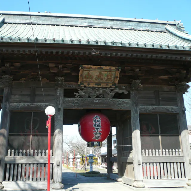 Daishoji Temple, Saitama A traditional Japanese temple gate with a large red lantern hanging in the center, intricate wooden carvings, and a tiled roof. Trees and buildings are visible in the background under a clear blue sky.