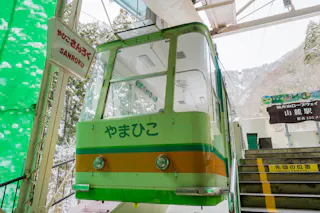 A green and white cable car named “やまひこ” waits at a snowy mountain station near stairs, with “SANROKU” signage on the left and snow-covered trees visible outside.