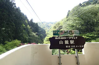 A brown sign with Japanese text stands on a walkway, with lush green trees and hills in the background. Overhead, cable car wires stretch towards the forested mountains under a bright sky.