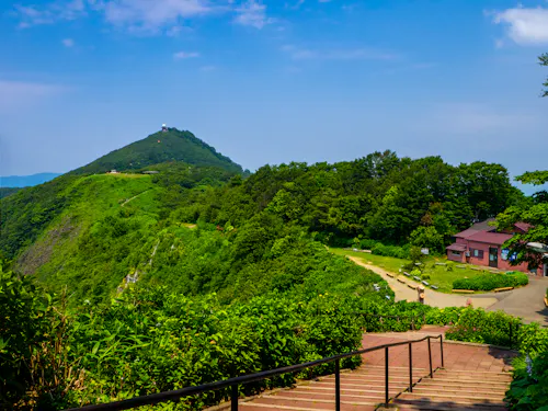 A scenic view of a lush green mountain under a blue sky, with a path and steps leading towards trees and a small building on the right. Shrubs and vibrant greenery surround the area.