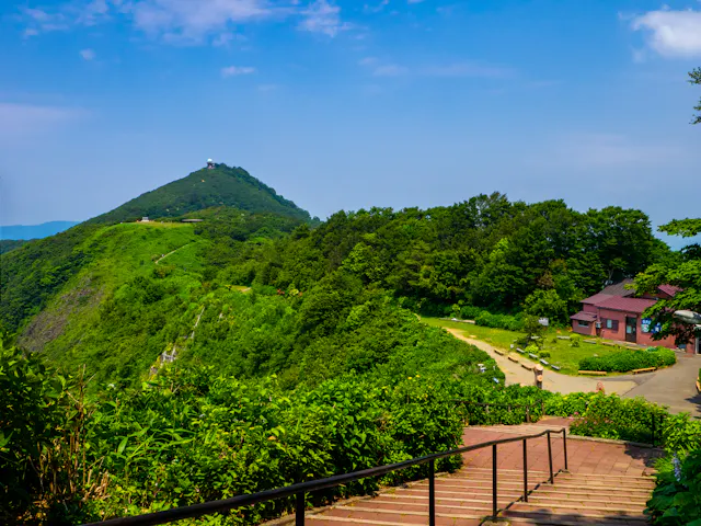 A scenic view of a lush green mountain under a blue sky, with a path and steps leading towards trees and a small building on the right. Shrubs and vibrant greenery surround the area.