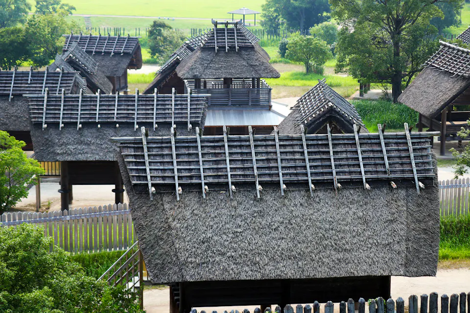 A collection of traditional thatched-roof buildings is surrounded by lush greenery. The structures feature wooden beams, and the scene has a rustic, historical feel. The background includes trees and a grassy field, creating a serene, nature-filled environment.
