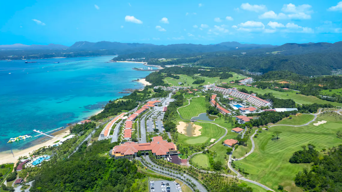Aerial view of the Kanucha resort in Okinawa, with the golf course, hotel buildings and a white-sand beach beside turquoise water