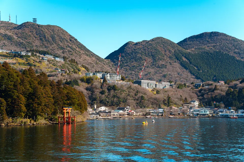 The red torii gate of Hakone Shrine standing in Lake Ashi near Moto-Hakone, with wooded hills and small buildings along the shore