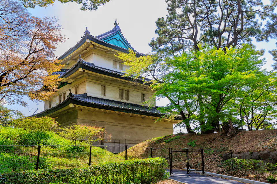 A white three-story Edo-period watchtower with a green roof stands on a stone base among spring trees in the Imperial Palace grounds