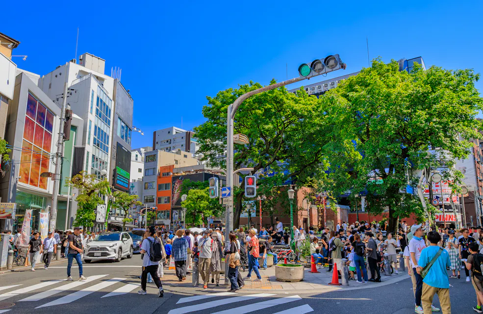 Amemura A busy city street intersection with many people walking and gathering, surrounded by tall buildings, green trees, cars, and clear blue sky on a sunny day. Traffic lights and crosswalks are visible.