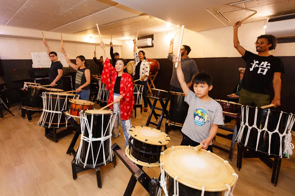 Adultos y niños levantando baquetas sobre tambores taiko en un taller