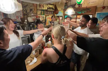 Travellers and local Osaka regulars raising sake cups in a lively toast inside a cluttered Namba izakaya on a bar-hopping night tour