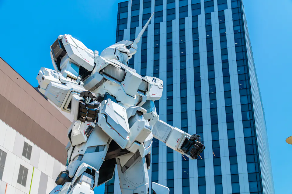The life-size Unicorn Gundam statue in Odaiba is a short Yurikamome ride from Tokyo Big Sight. The life-size white Unicorn Gundam statue in Odaiba posed in front of a tall glass building under a blue sky