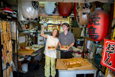 Two tour guests holding sushi boxes among red paper lanterns at a Tsukiji shop in Tokyo