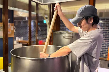 Brewer stirring mash in a large stainless steel kettle with a wooden paddle at an Osaka brewery