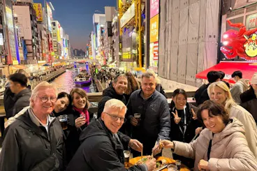 Tour group enjoying food and drinks beside the neon-lit Dotonbori canal on an Osaka Namba evening food tour